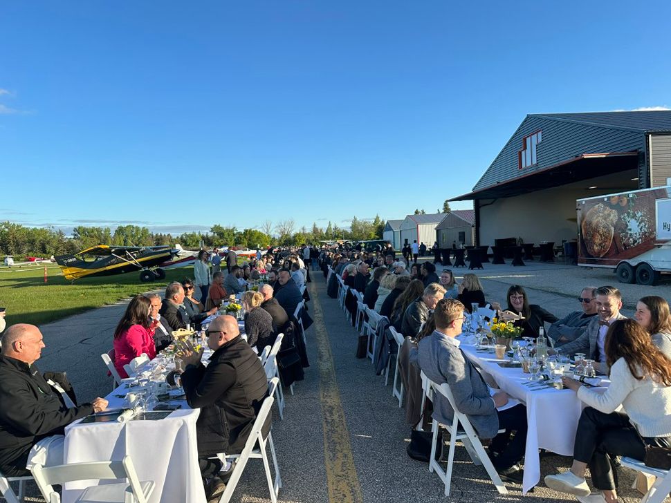 Two rows of long tables with white tablecloths and flowers in vases, food, and people seated at the tables.