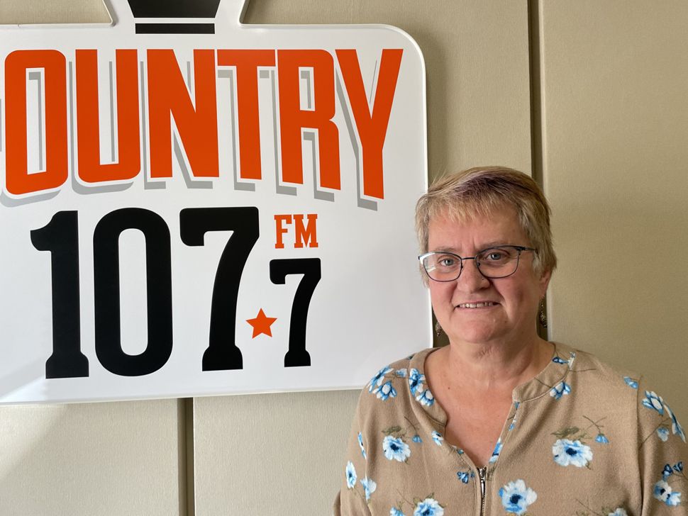 A woman with short blond hair smiles while standing near the Country 107 sign.