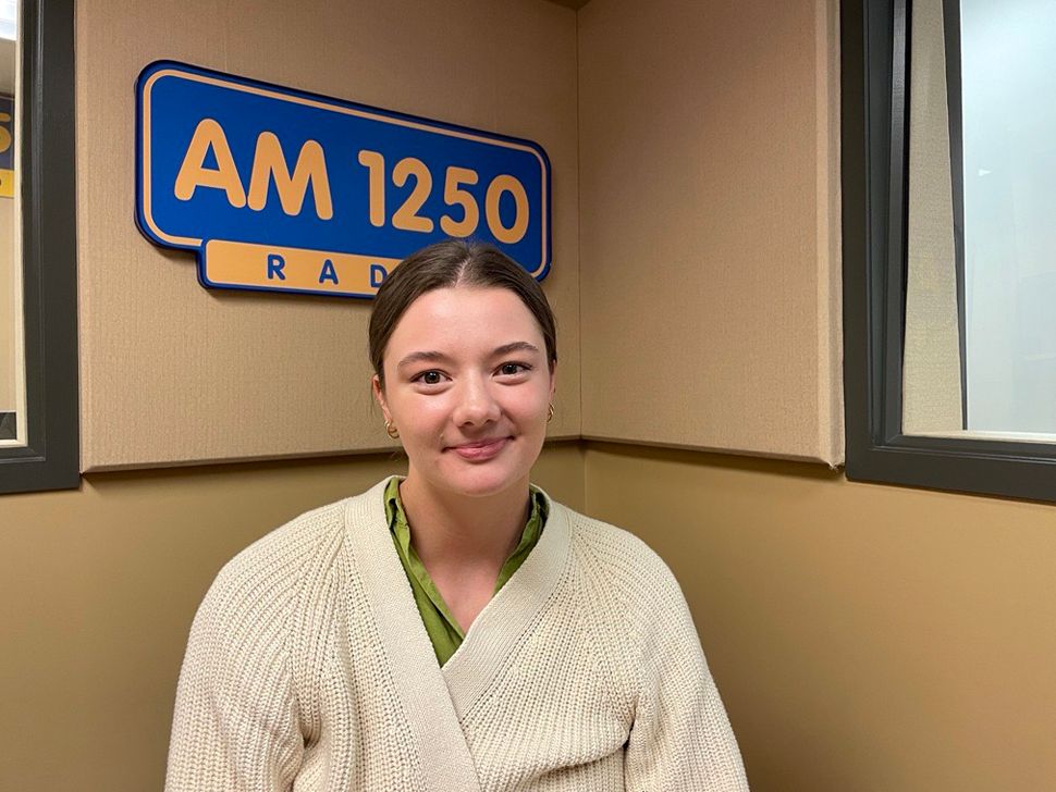 Une jeune femme souriante. Il y a un panneau radio AM1250 derrière elle.