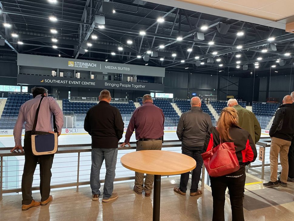 Six men and one woman look over a railing down to the ice surface of an arena.