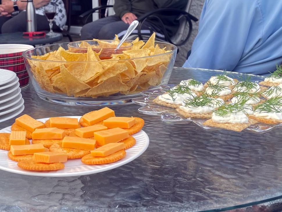 A glass patio table with a stack of plates near larger plates with snacks like crackers and cheese, chips and dip.