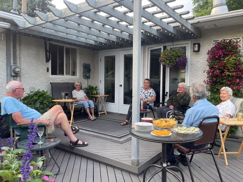 Two men and four women sitting on a deck in a circle-type style. There is a table with snacks on it.