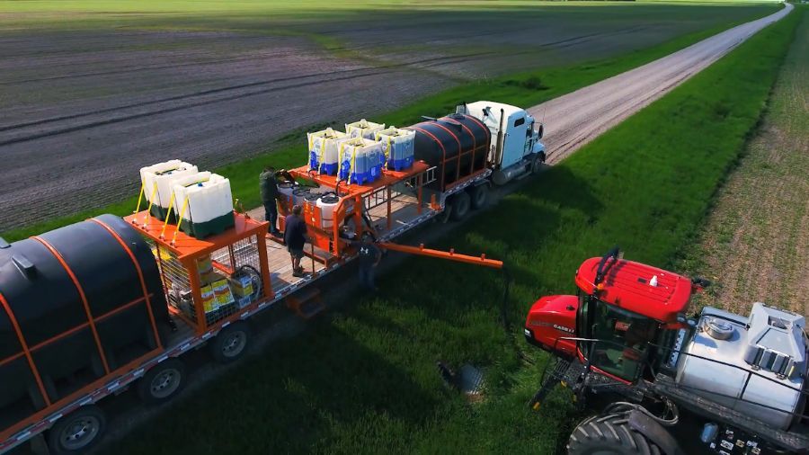 a flat deck trailer with massive plastic tanks and a field sprayer drawing liquid from them