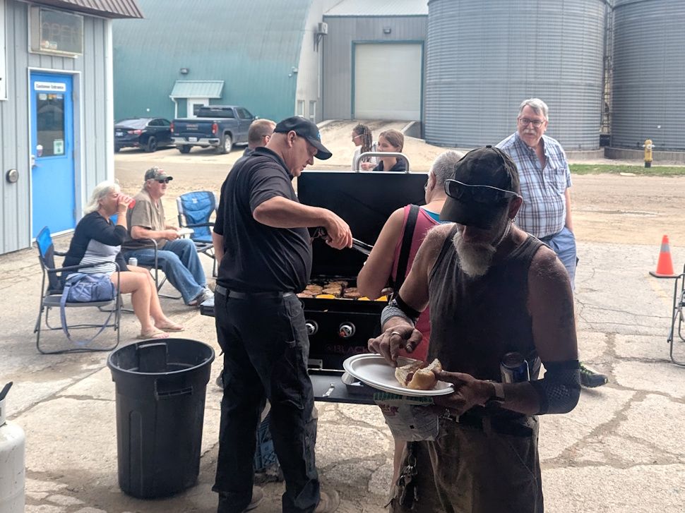 People gathered outdoors enjoying BBQ food at a fundraiser event, with plates of burgers, hot dogs, and drinks visible.