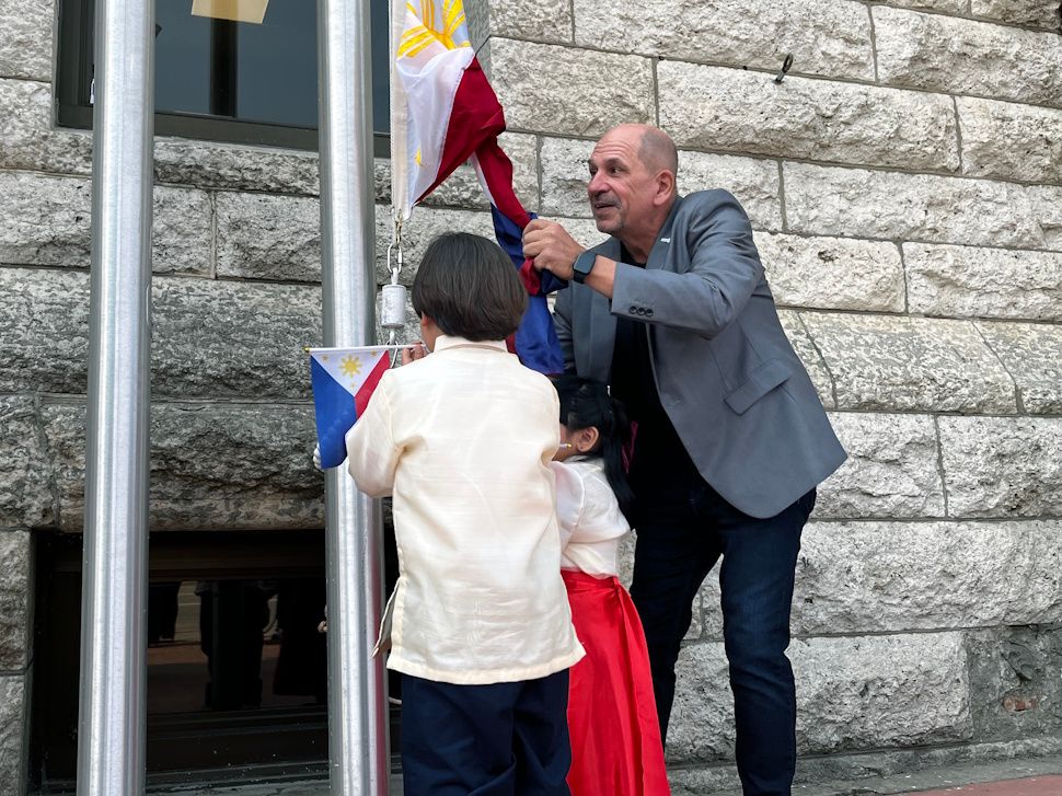 Deputy Mayor Faron Nicholls helps two children in traditional Filipino attire raise the Philippine flag during the ceremony at City Hall.